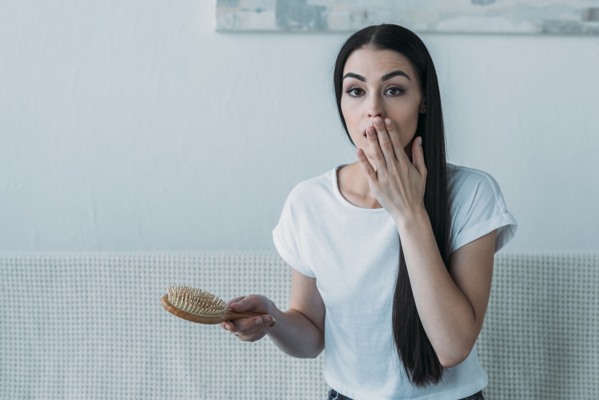 shocked brunette woman holding hairbrush and looking at camera, hair loss concept